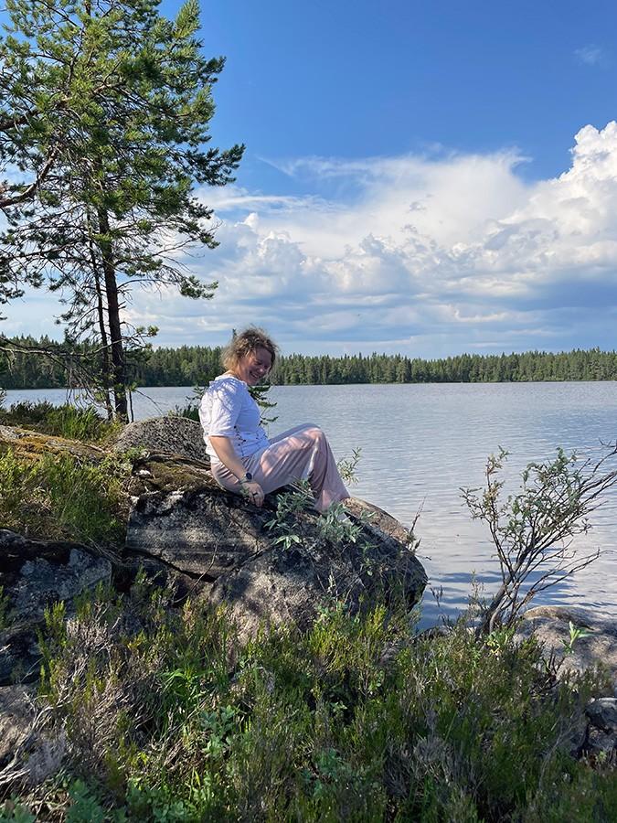 Nainen istuu kivellä, järven rannalla ja hymyilee.