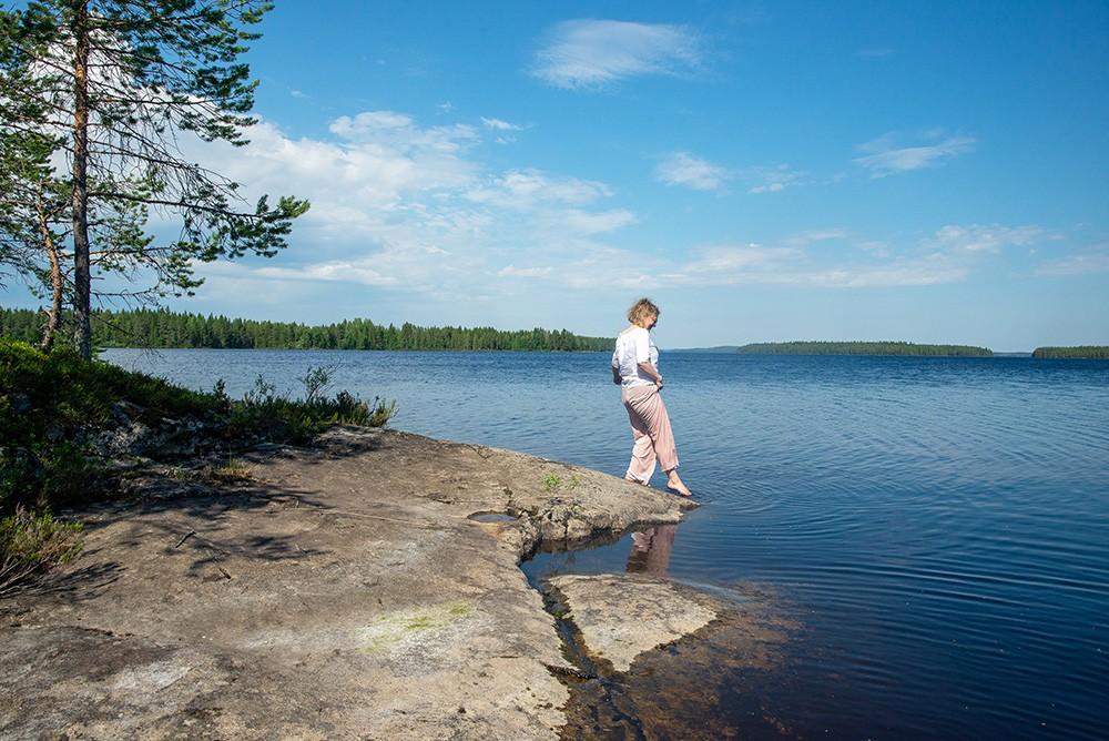 Nainen seisoo järven rannalla ja koskettaa varpaillaan vettä.