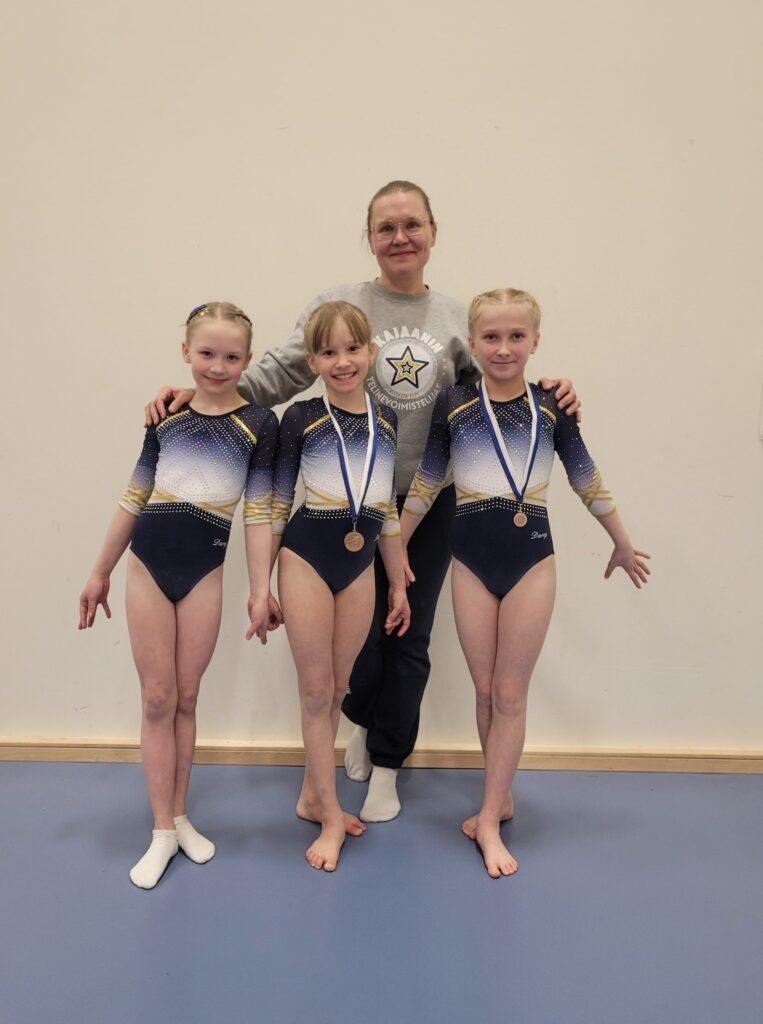 A woman stands behind three young gymnasts. They are all looking at the camera and smiling.