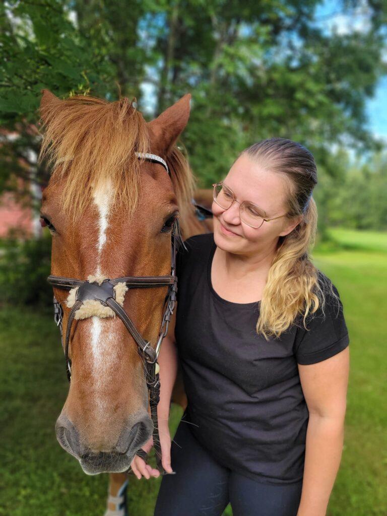 Nainen hymyilee hevosen vieressä ja katsoo hevosta.